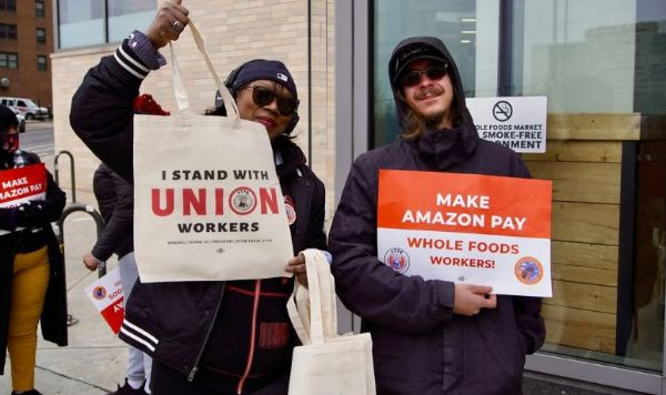 Rally attendees hold a tote bag that reads, “I stand with union workers” and a sign that reads, “Make Amazon pay Whole Foods workers!”