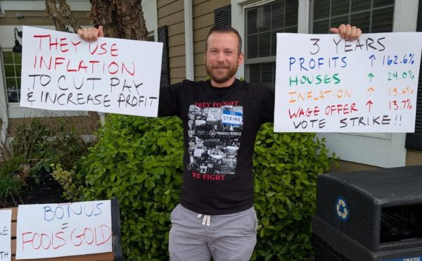 A person holds a sign showing Pratt & Whitney profits have increased over three years while wages haven’t kept up with the cost of living, and another that reads, “They use inflation to cut pay & increase profit.”