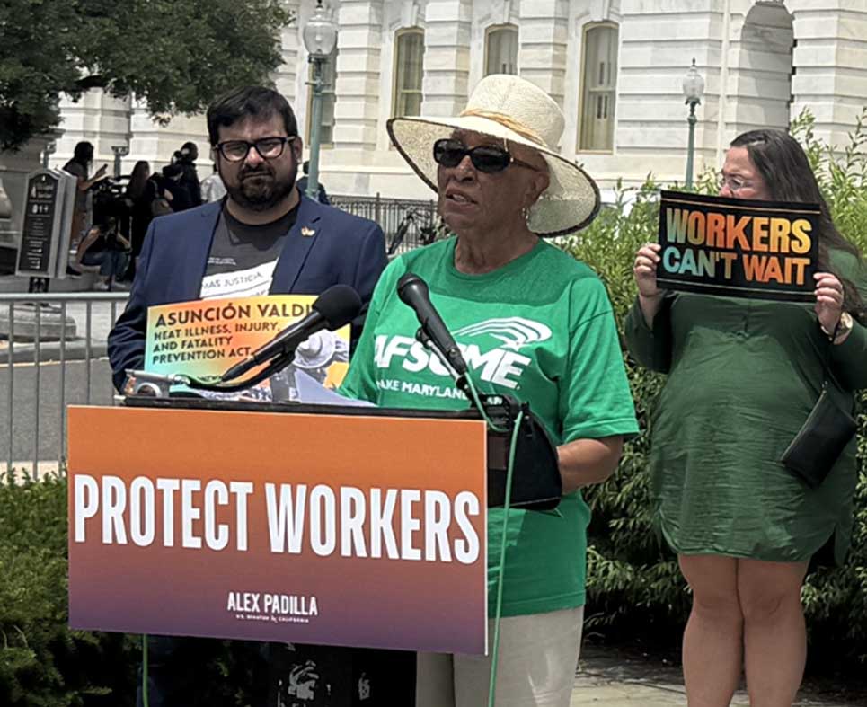 A woman in an AFSCME tshirt speaking at a heat rally with a sign that says 'Protect Workers'